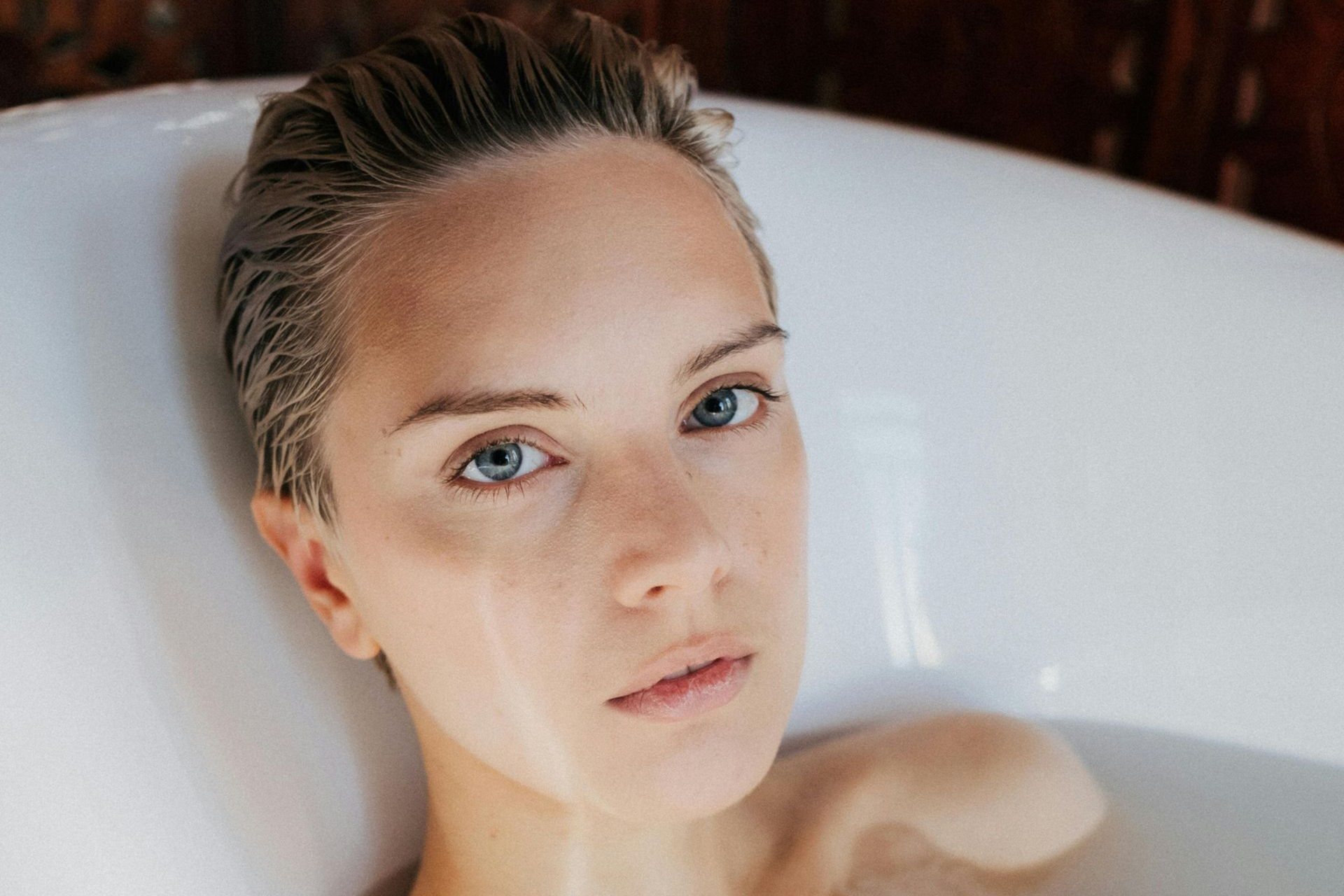 Woman with short, wet hair reclines in a white bathtub, looking at the viewer.