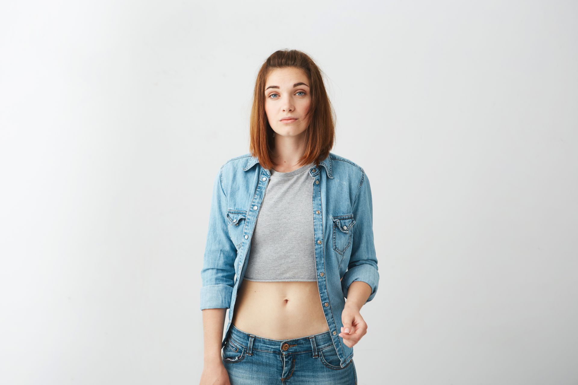 A woman in a crop top and jeans is standing in front of a white wall.