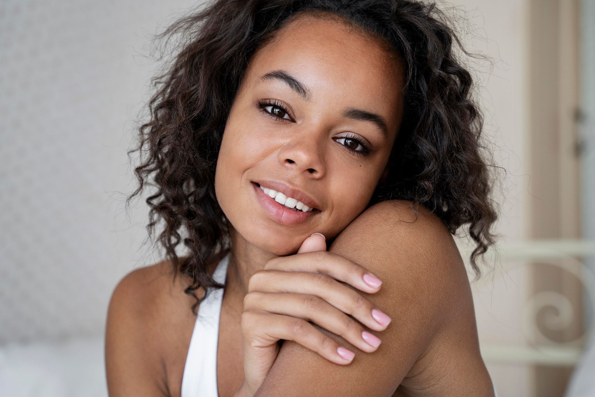 A woman with curly hair is smiling with her hands on her shoulder.