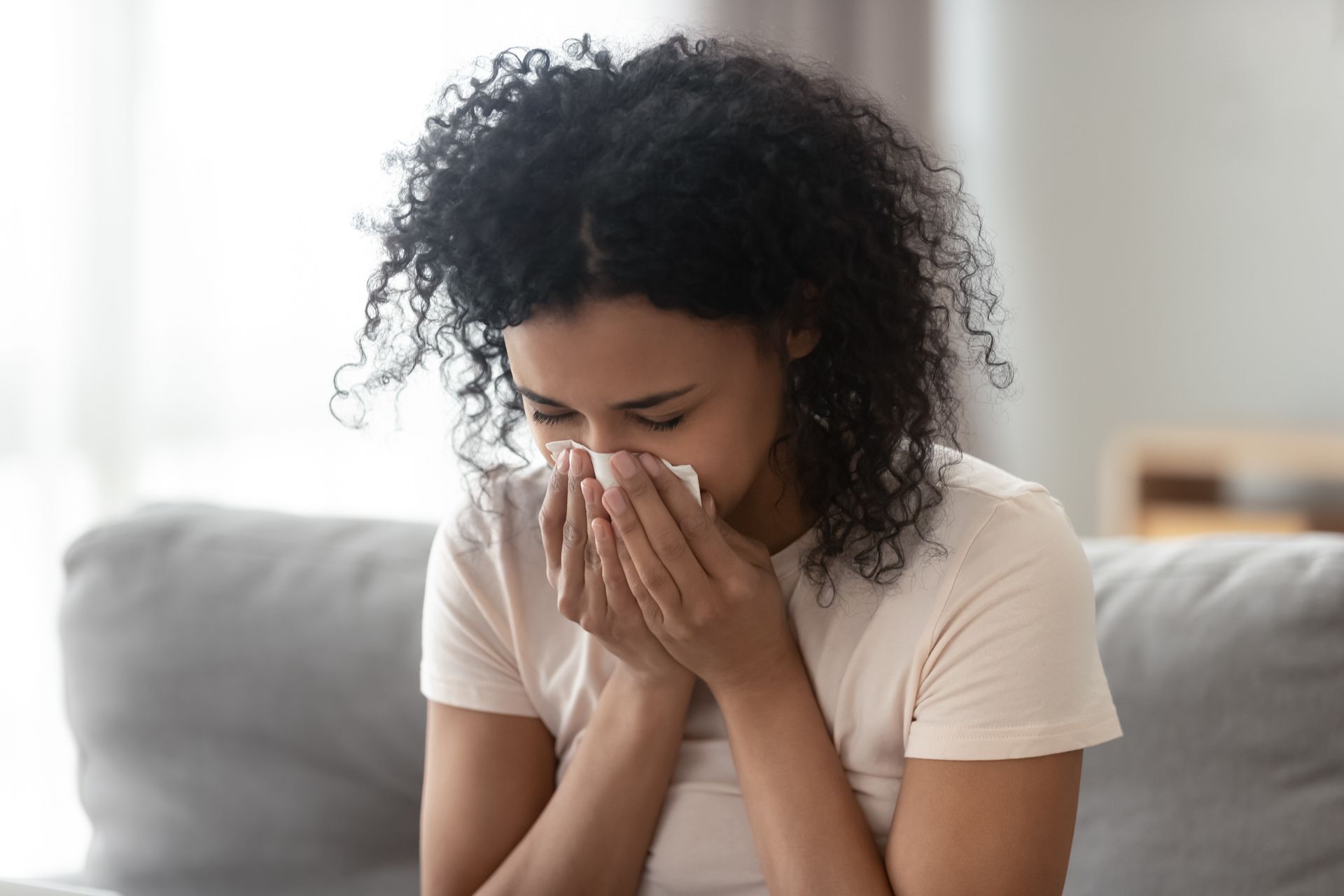 A woman is blowing her nose into a napkin while sitting on a couch.