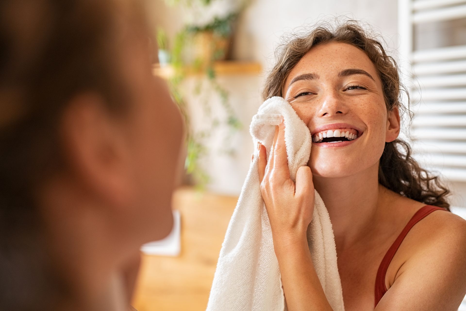 A woman is smiling while cleaning her face with a towel in front of a mirror.