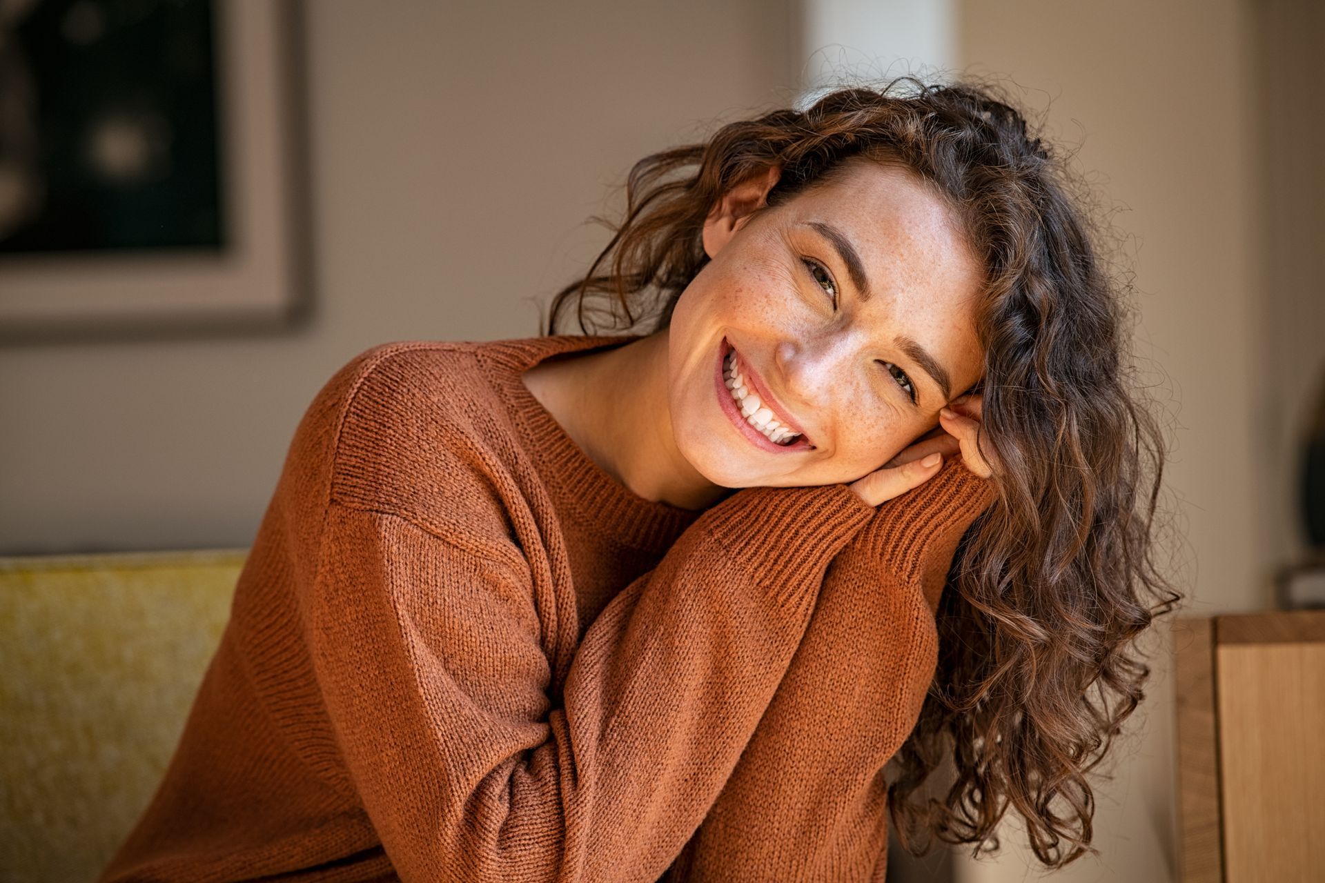 A woman in a brown sweater is smiling while sitting on a couch.