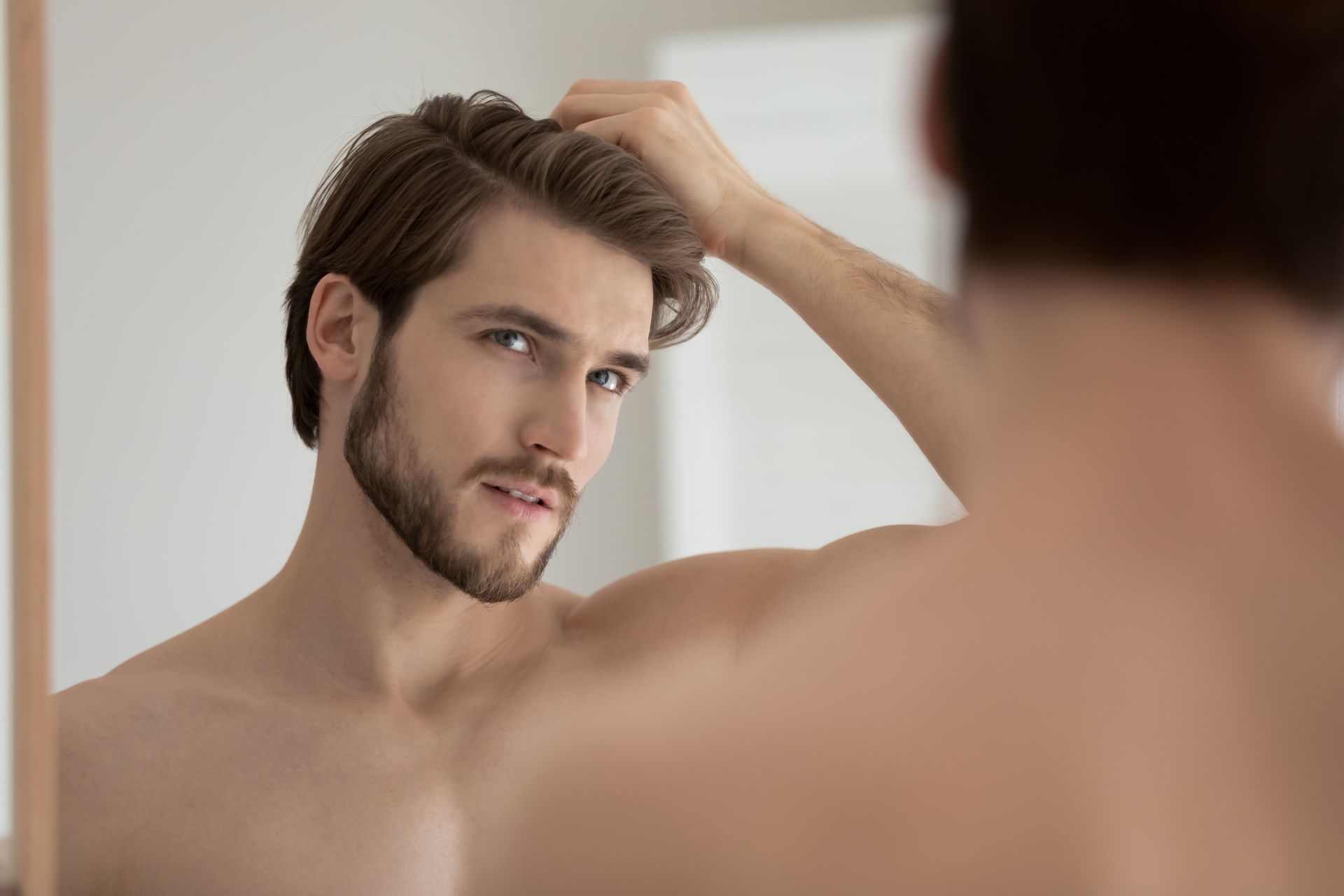 A shirtless man with a beard is looking at his hair in the mirror.