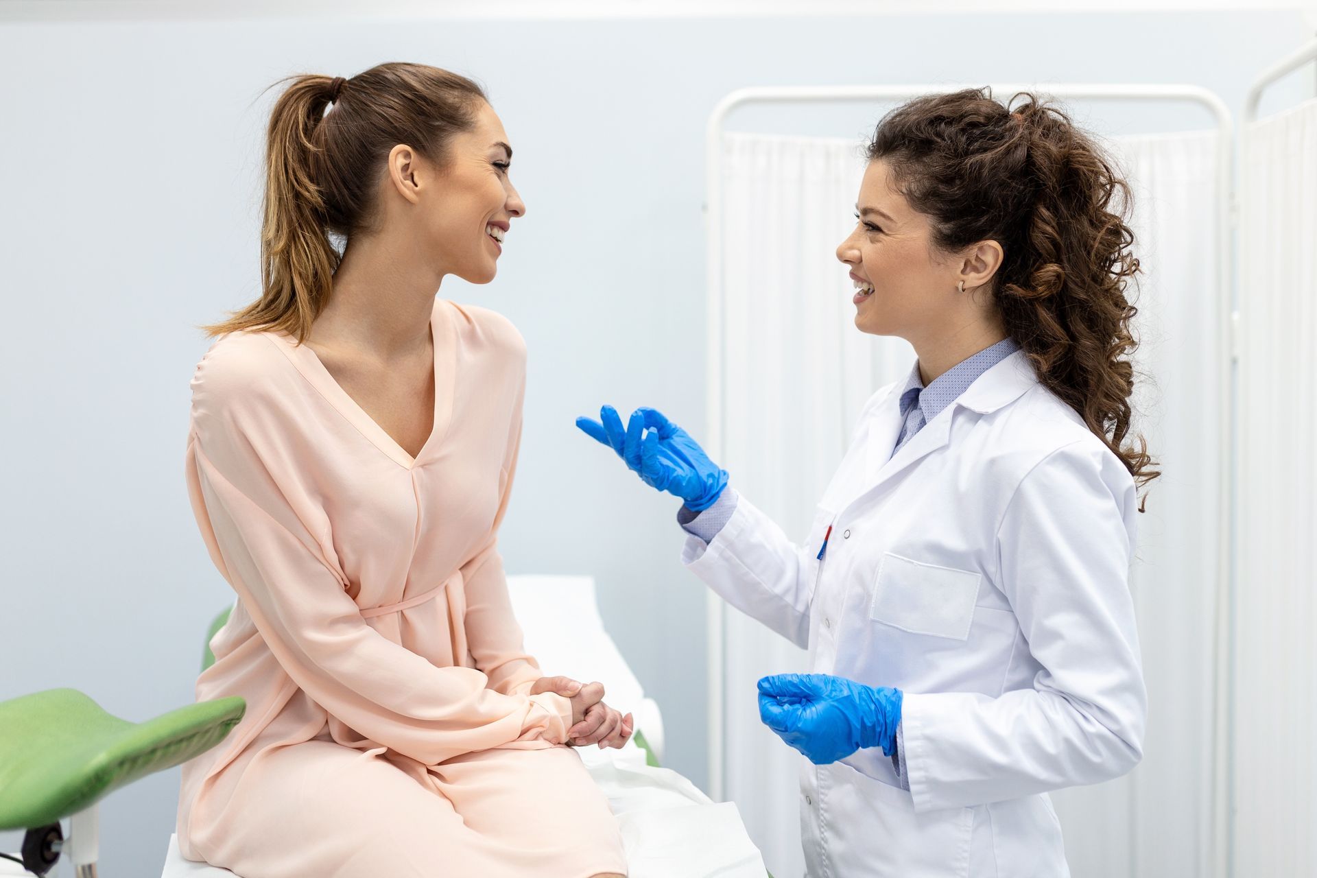 A doctor in blue gloves talks to a patient, in a medical exam room.