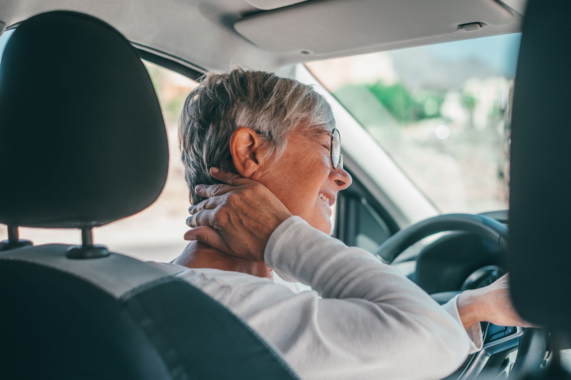 Woman in a car, hand on neck, grimacing. Driving with window in the background.
