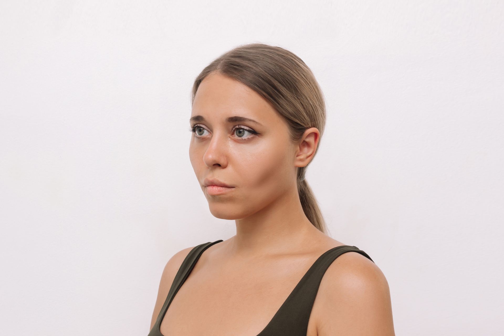 Woman with light brown hair in a low ponytail, wearing a dark green tank top, looking to the side with a serious expression.