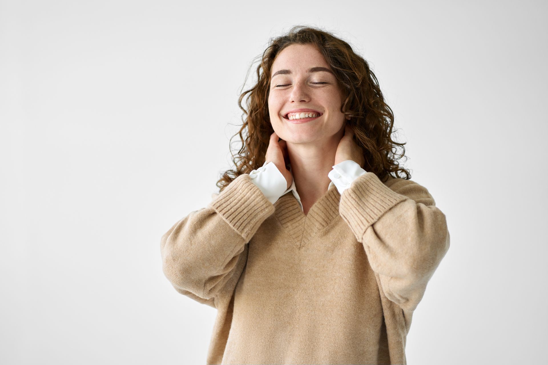 A woman in a tan sweater is smiling and touching her neck.