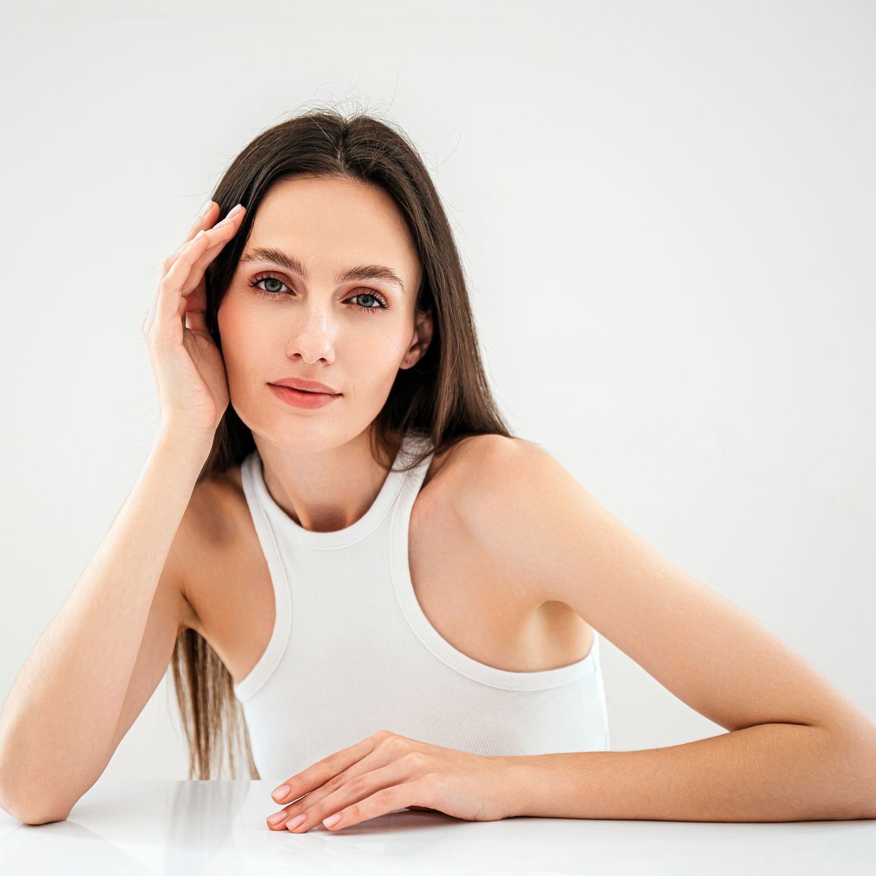 A woman in a white tank top is sitting at a table with her hand on her face.