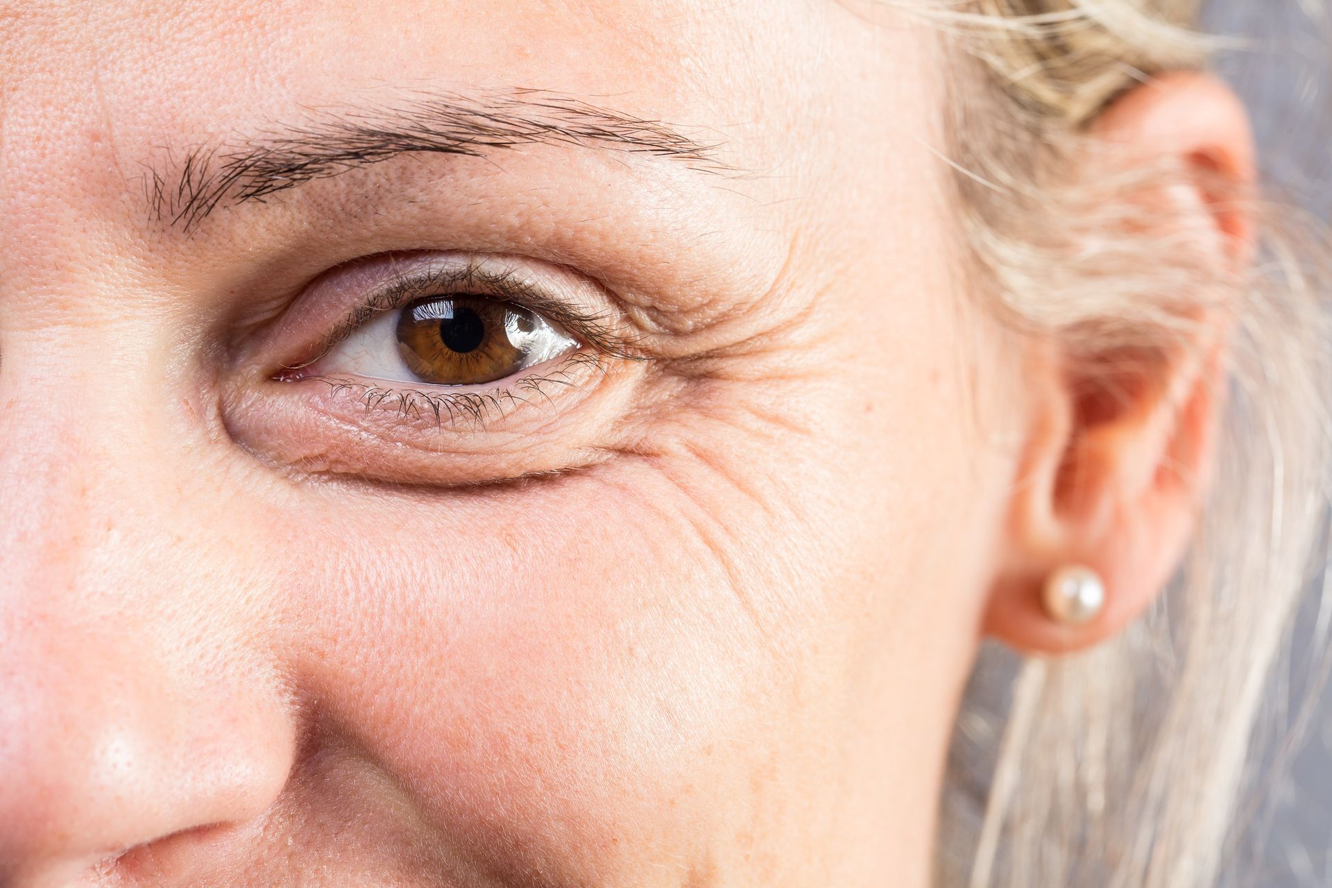 Close-up of a person's eye, showing brown iris, with wrinkles around the eye and a pearl earring.