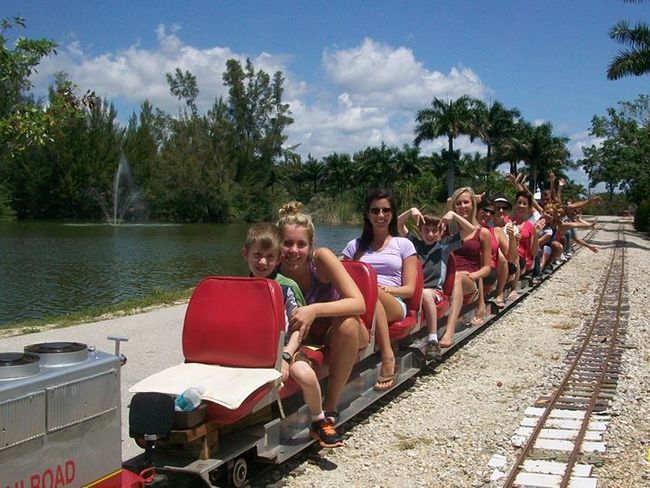 People ride a small train on tracks beside a lake with a fountain. The sky is blue with some clouds.