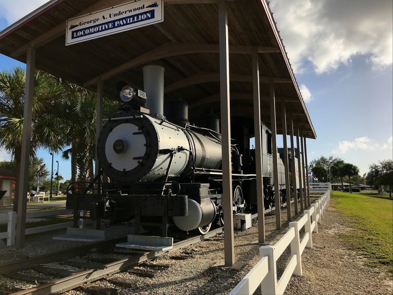A vintage gray steam train under a wooden shelter, with a white picket fence alongside a grassy area.