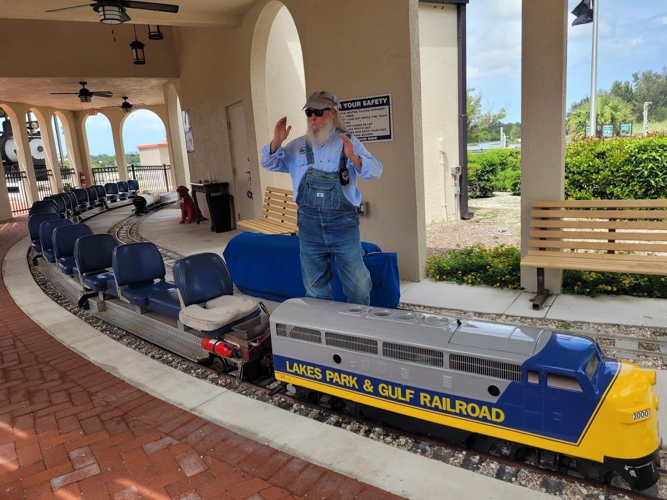 A man in overalls waves beside a small blue and yellow train at the Lakes Park & Gulf Railroad station.