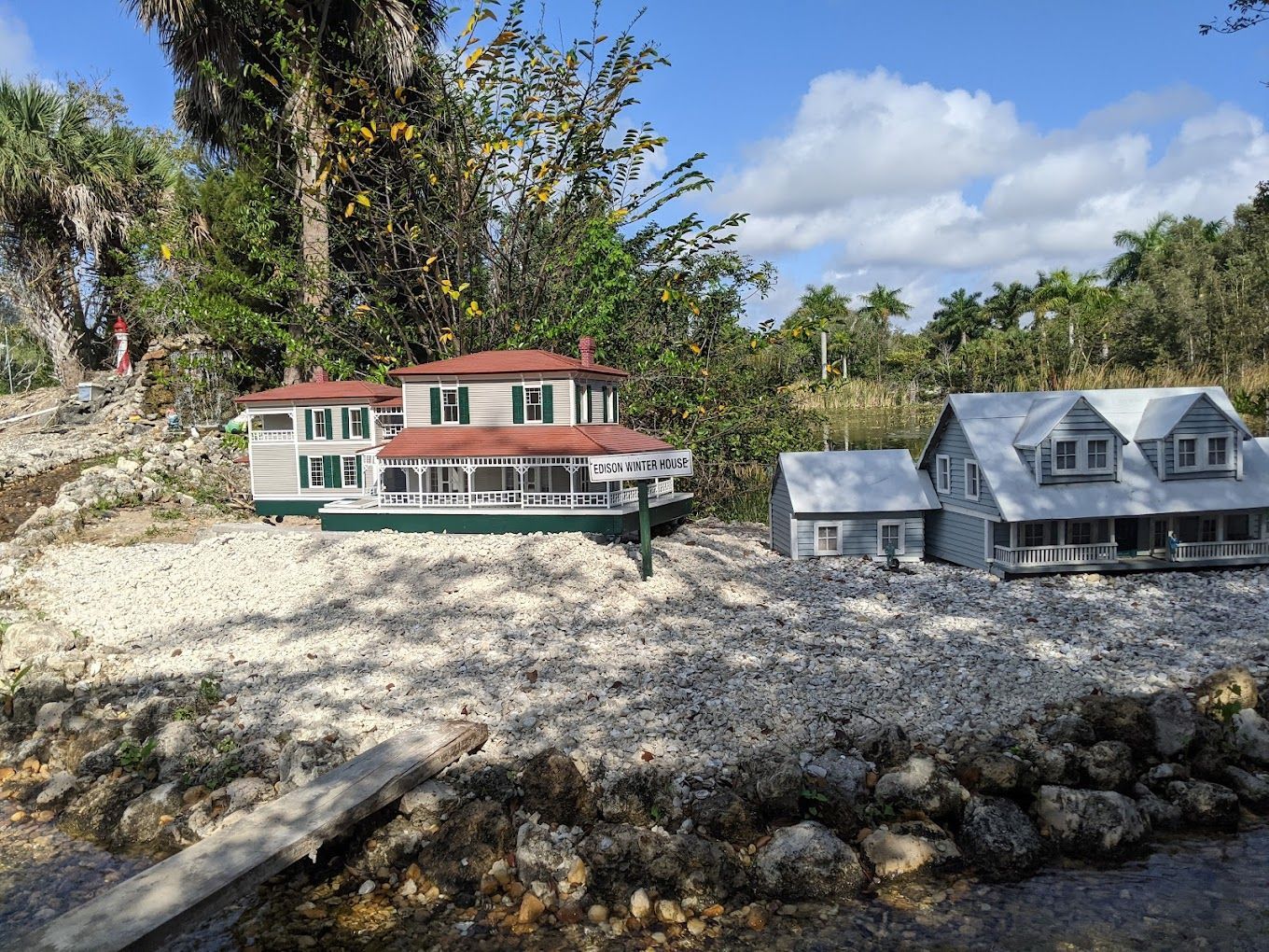 Miniature houses with white and green trim sit on a bed of gravel under a sunny sky, with trees in the background.