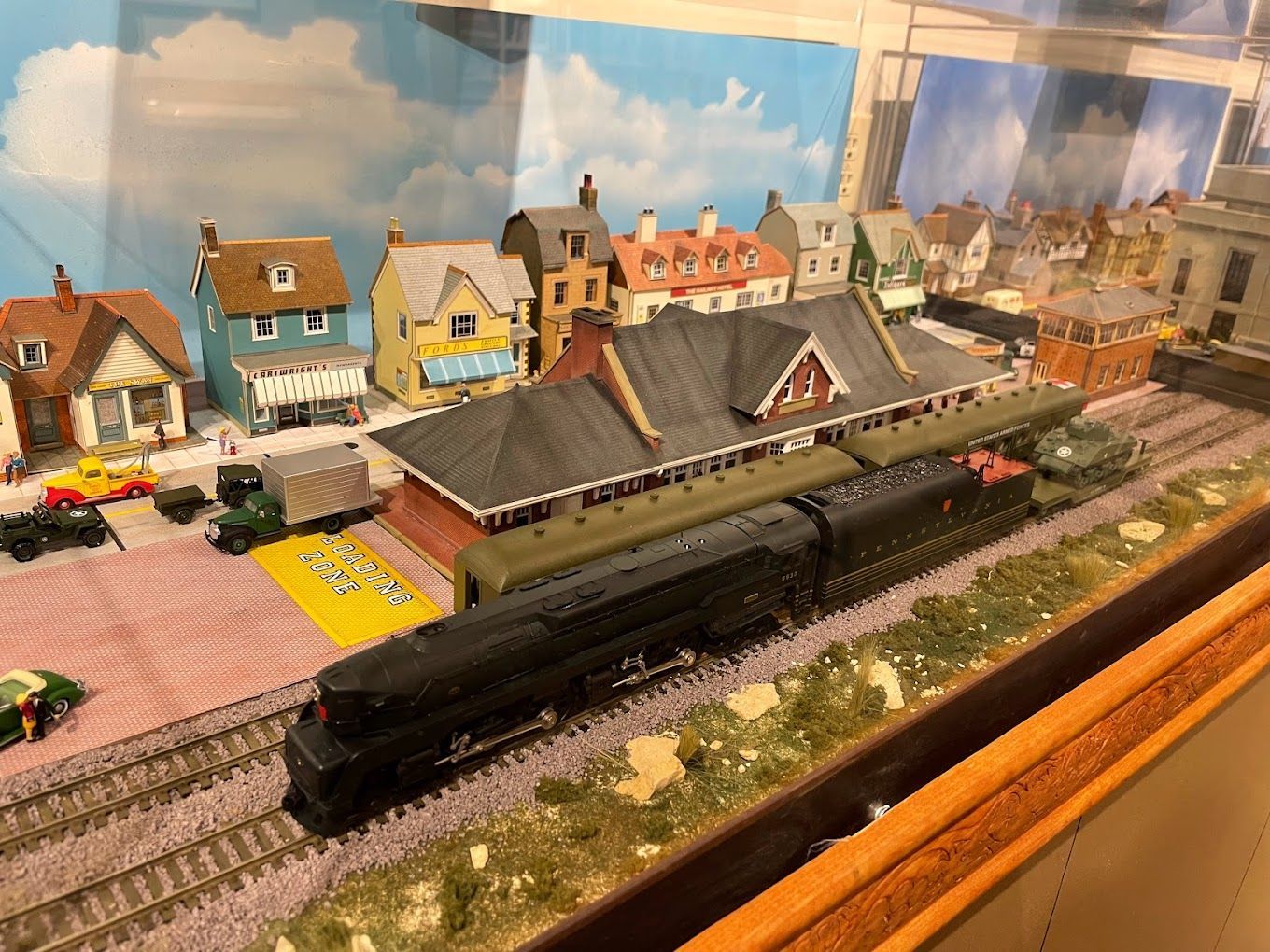 Model train station scene with a black locomotive, passenger cars, and colorful buildings in the background.