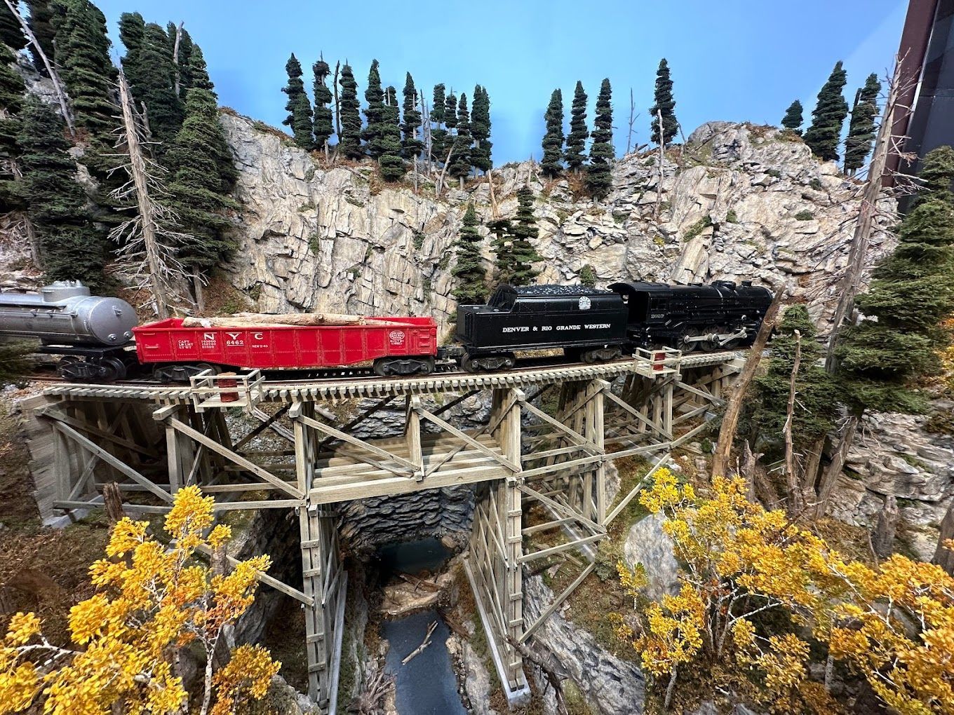 Model train crossing a wooden trestle bridge over a tunnel and river, surrounded by rocky terrain and trees. The train is red, black, and silver.