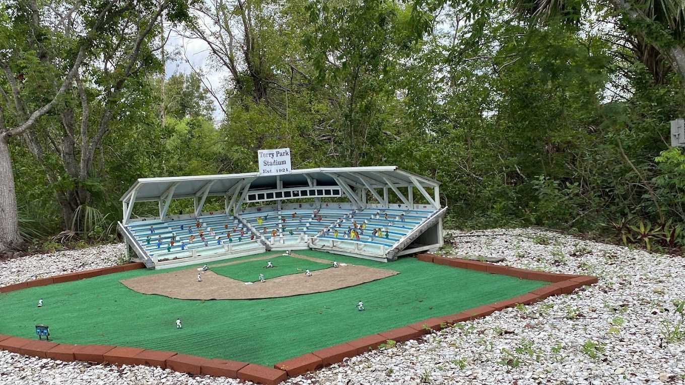 Miniature baseball stadium with green field and blue and white seating, set outdoors with trees and white rocks.