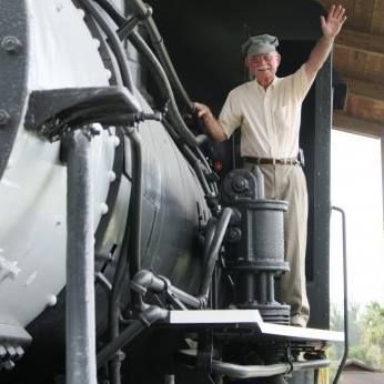 Man waves from the open cab of a black steam train. He wears a cap and tan clothing, smiling.