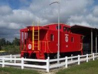 Red caboose with yellow accents behind a white fence and under a partly covered area.