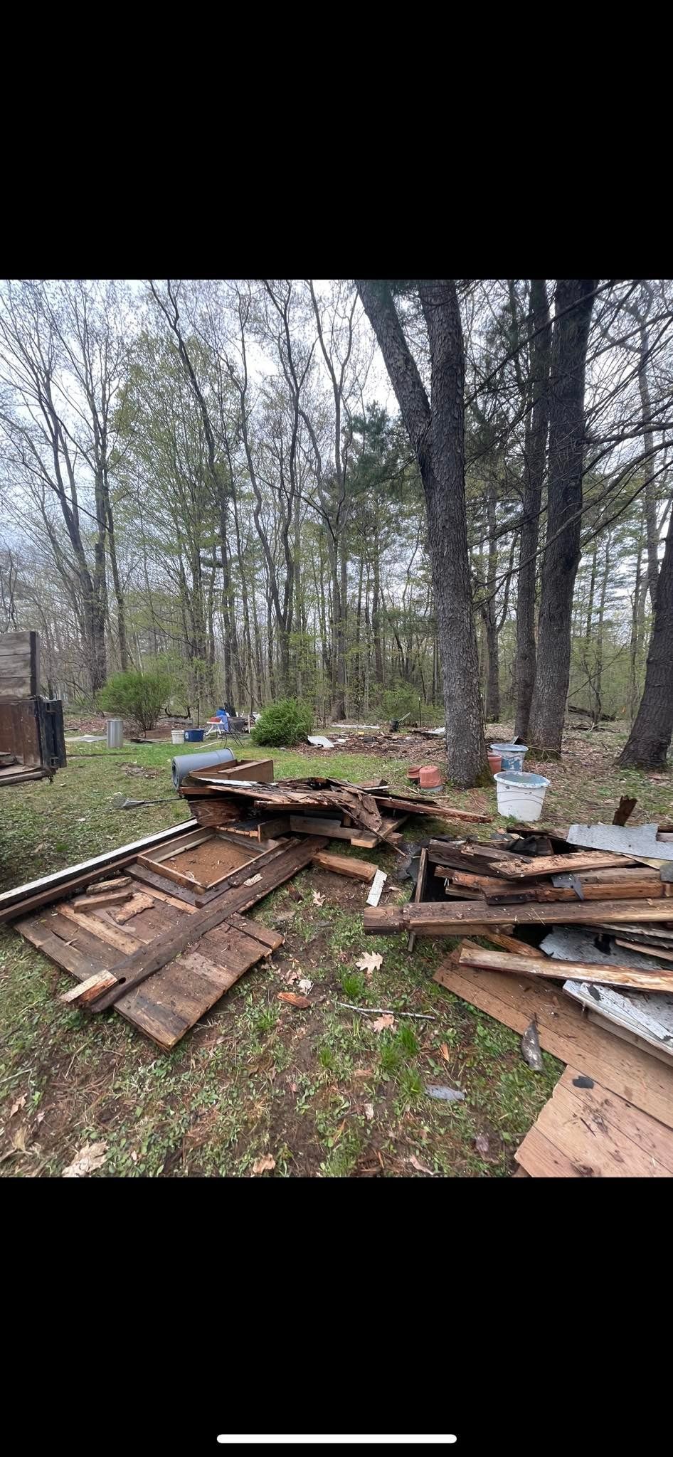 Debris and wood scraps from a dismantled structure scattered on the ground in a wooded area.