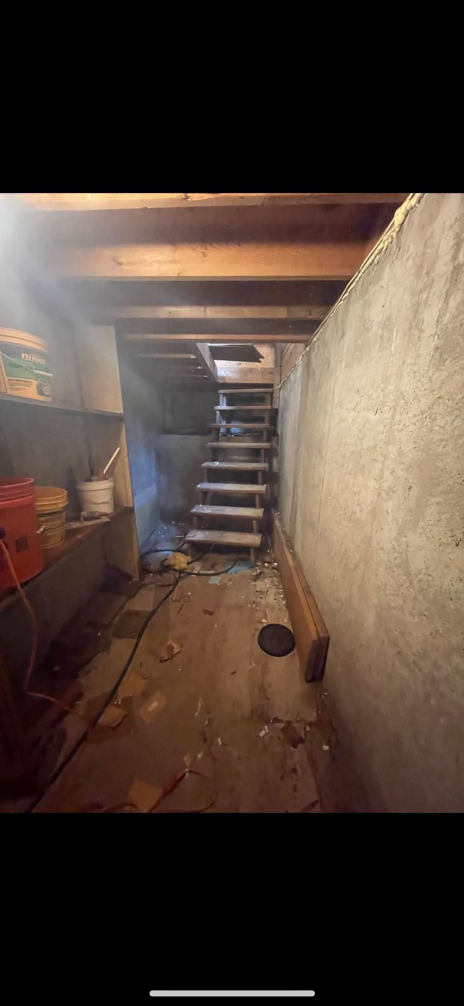 A dim basement hallway with a rough wall on the right, a cluttered workbench on the left, and stairs leading upward.