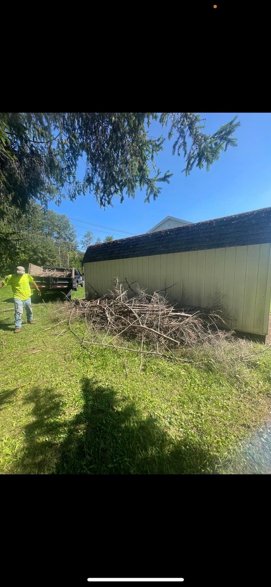 A person in a high-visibility vest clears a large pile of brush next to a long, light-colored fence on a sunny day.