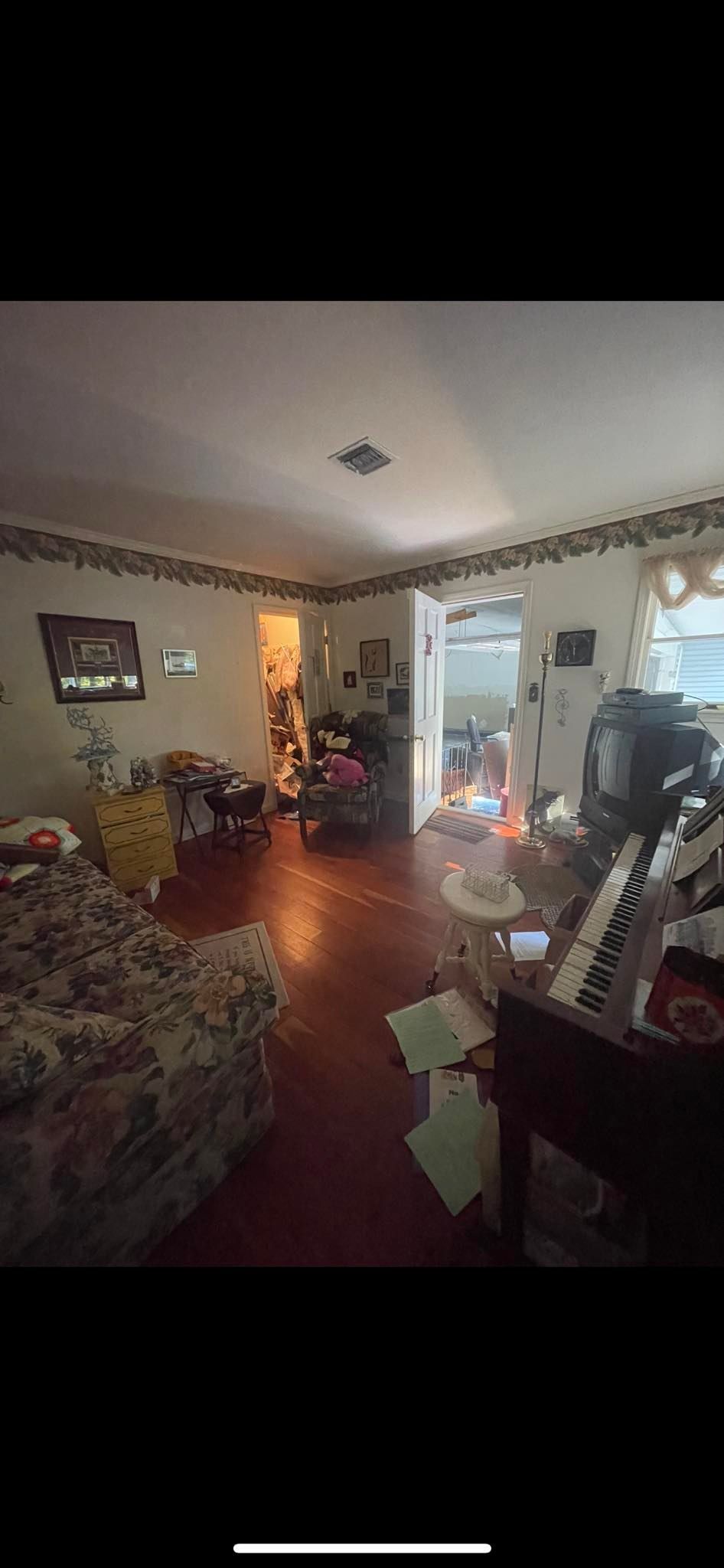 A dimly lit, cluttered living room with a patterned sofa, a piano, and a doorway leading to a brightly lit room.
