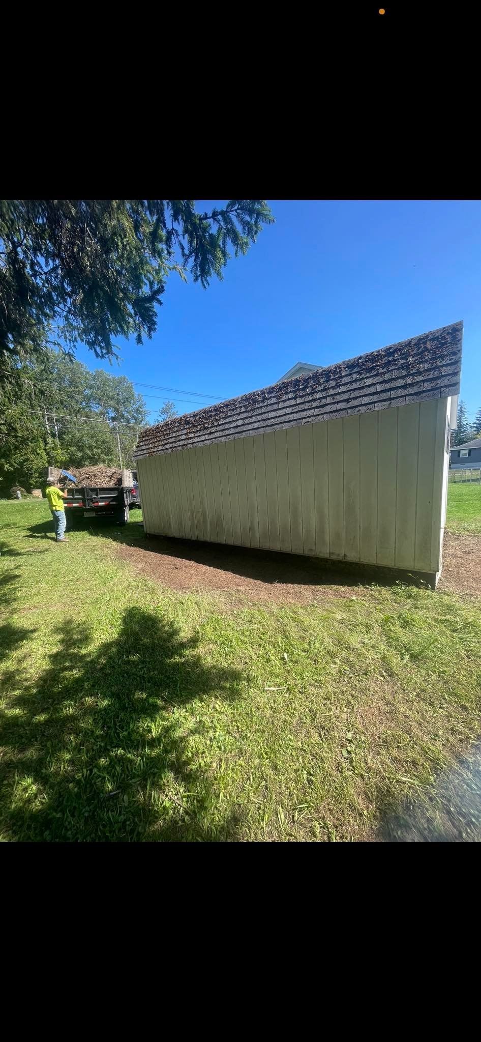 A white shed sits on a grassy lawn next to a parked trailer on a sunny day.