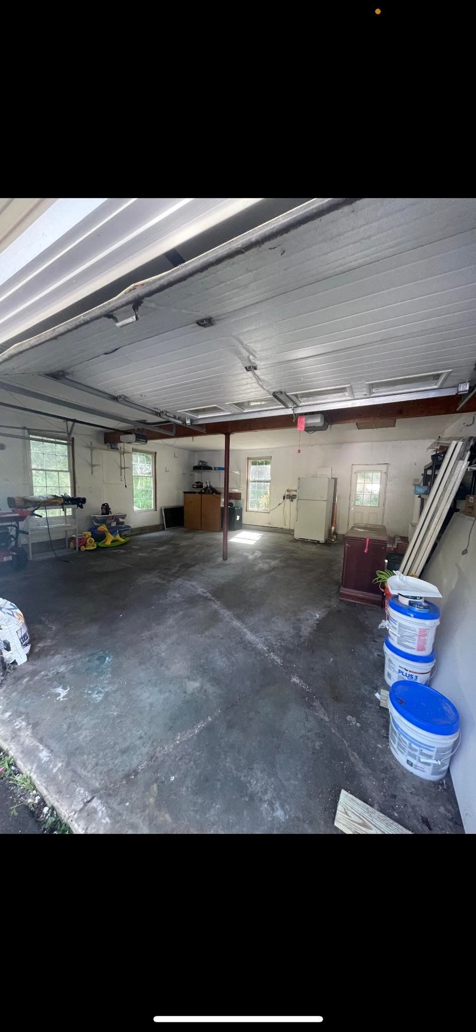 A view from the entrance of a garage, showing a concrete floor, white walls, several windows, and stored supplies.