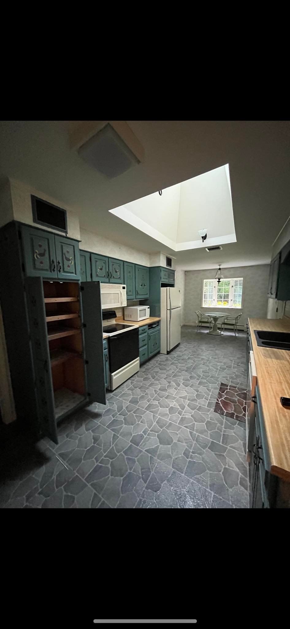 A kitchen with dark teal cabinets, gray tiled flooring, a skylight, and a dining area in the background.