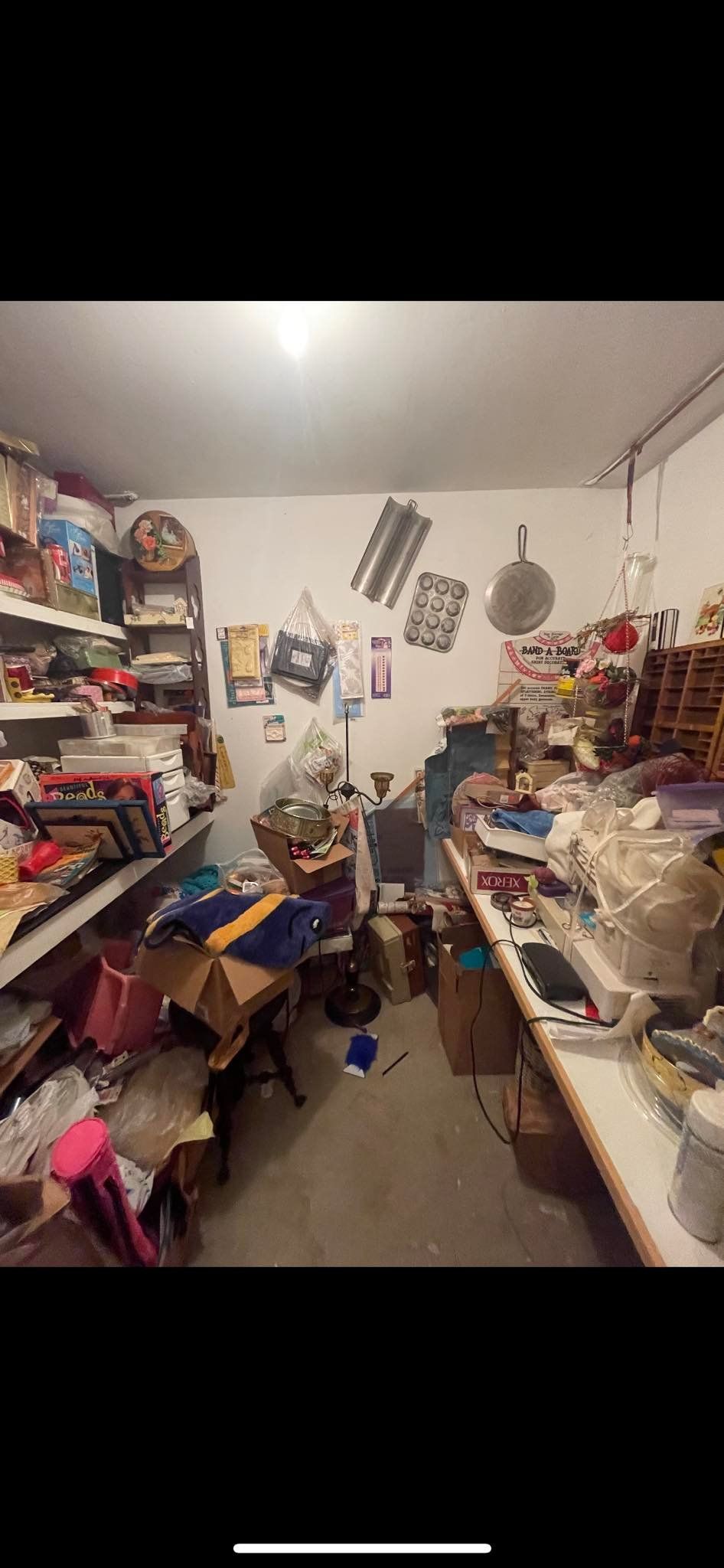 A cluttered storage room or garage with shelves packed with items, boxes on the floor, and various objects hanging on walls.