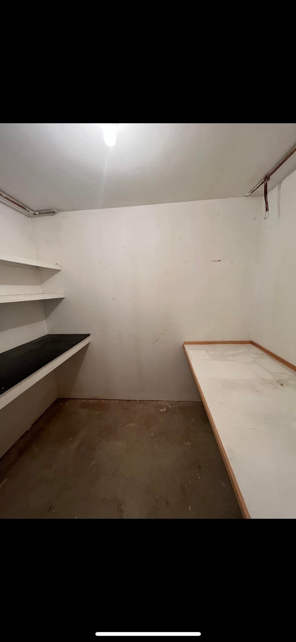 A narrow, minimalist storage room with white walls, a dark-topped shelf on the left, and a white counter on the right.