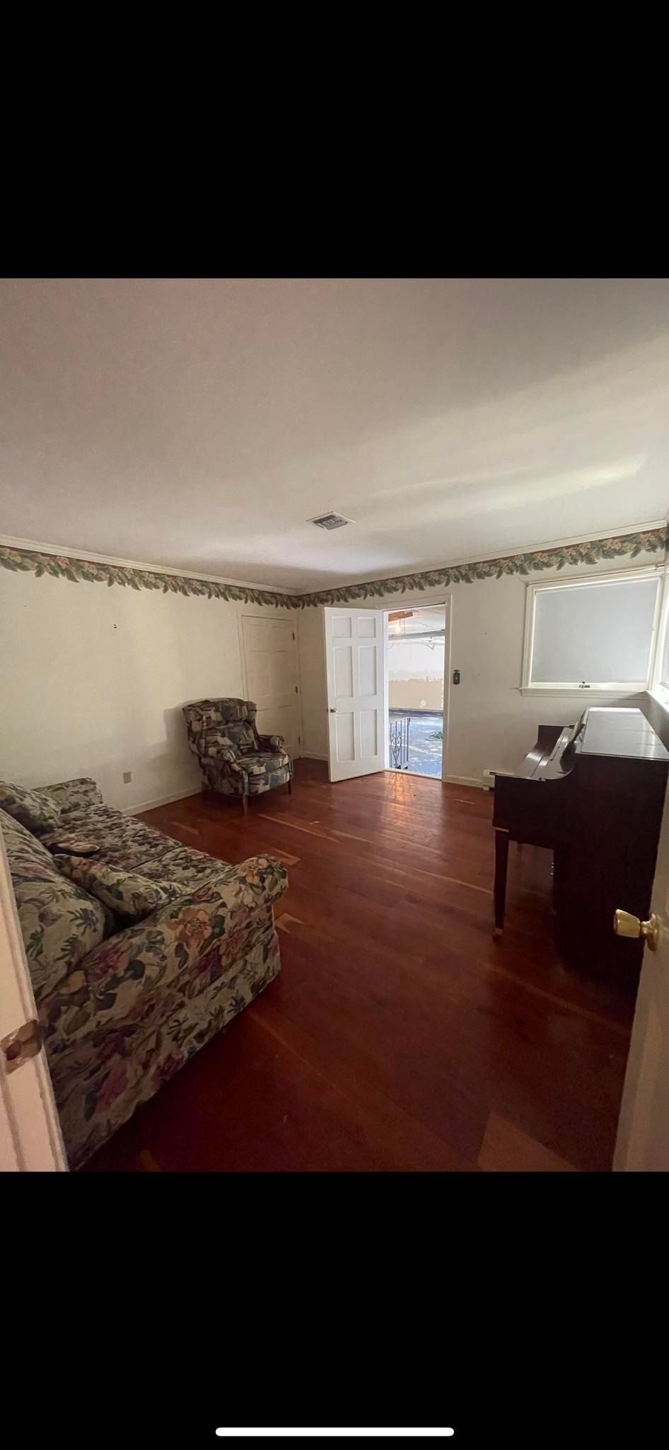 A living room with wooden floors, floral furniture, wallpaper border, and an open doorway leading to another room.