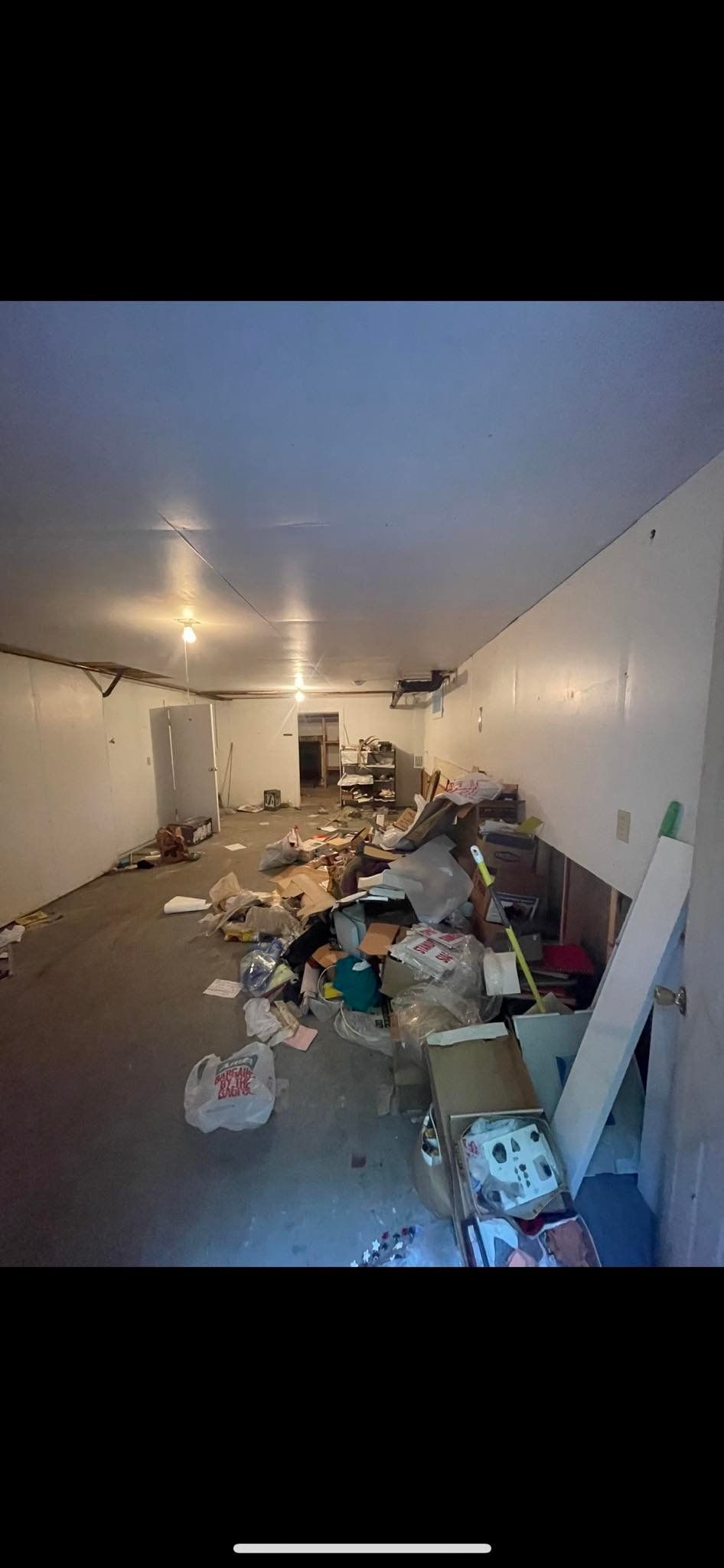 A long, narrow room with white walls and a concrete floor, scattered with trash, boxes, and miscellaneous debris.