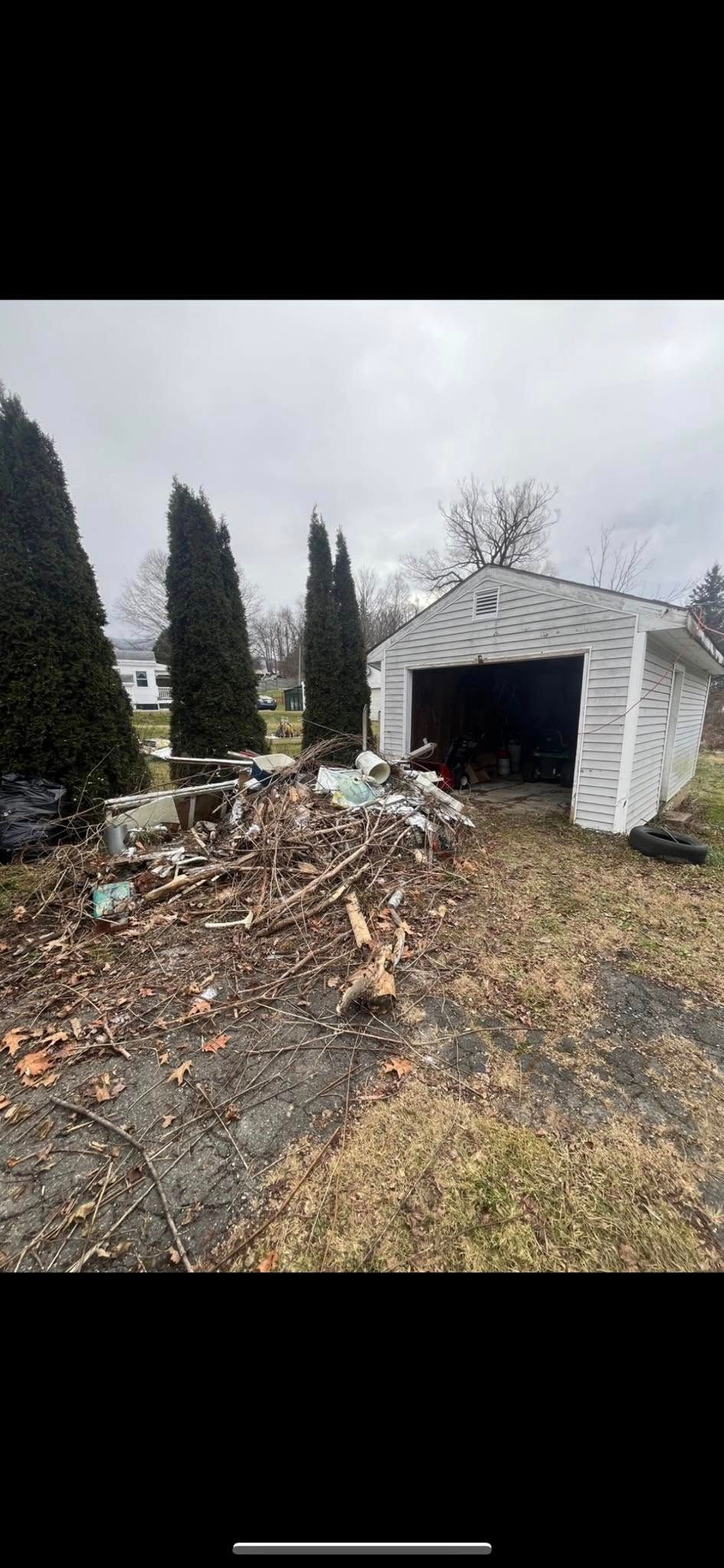 A pile of brush and debris sits in front of a small white garage on a cloudy day, flanked by three tall evergreen trees.