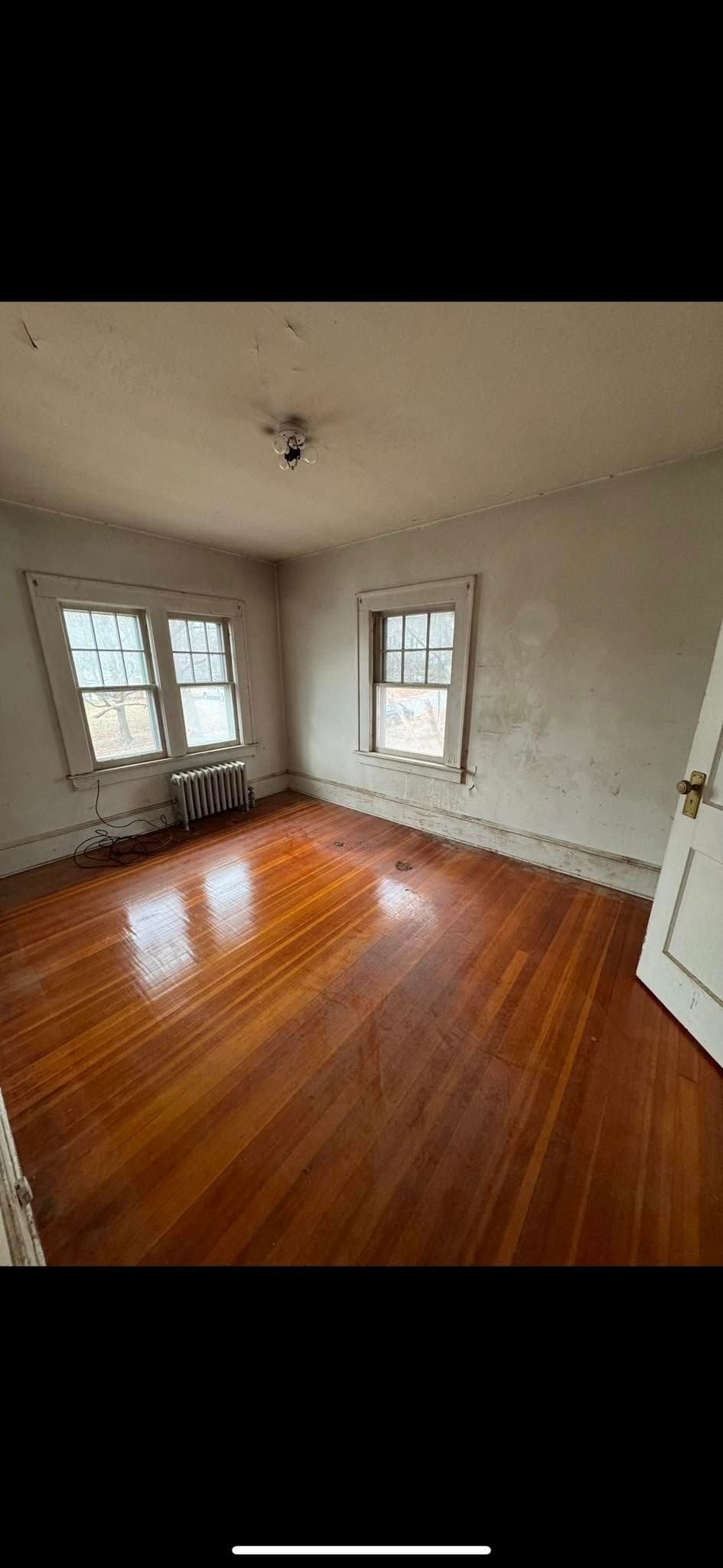 An empty, sunlit room with polished wood floors, plain walls, and two windows.