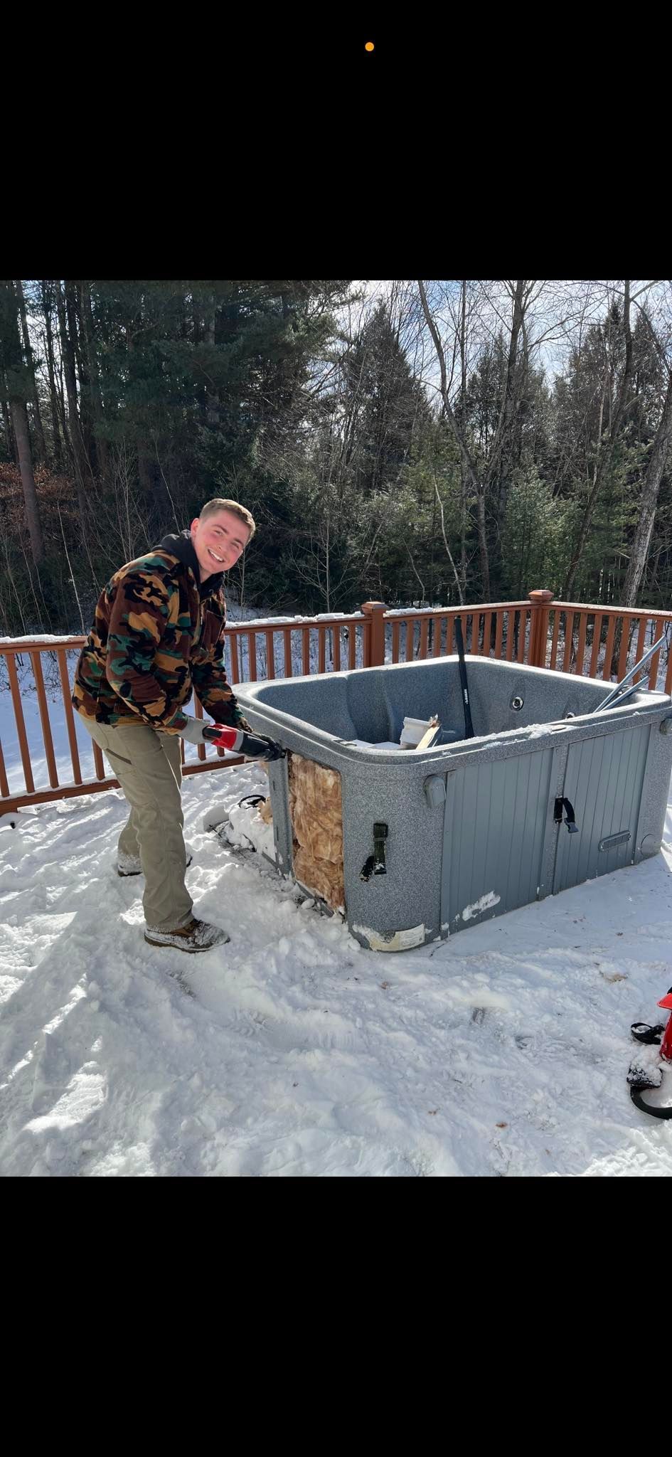 A person in a camouflage jacket works on a gray, square structure on a snow-covered deck with trees in the background.