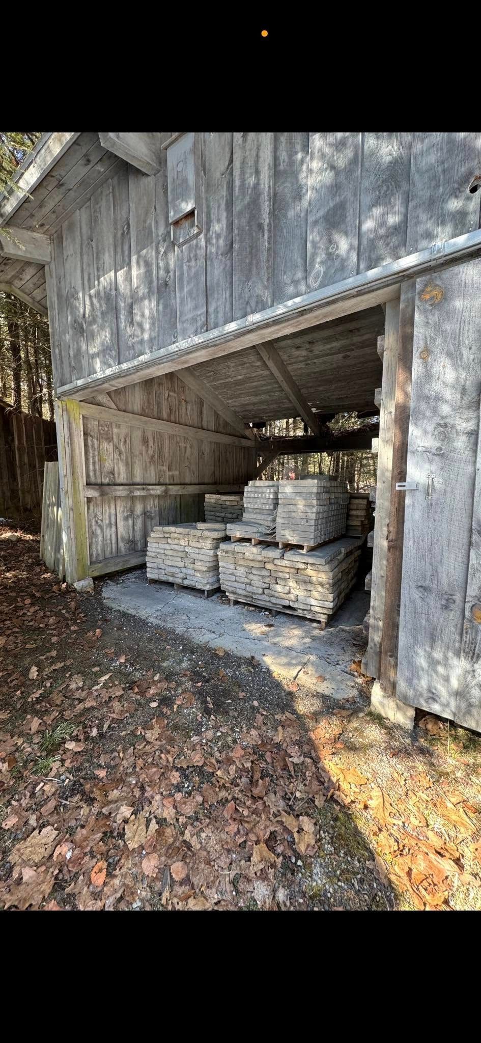 Stacks of grey construction materials stored under the open, wooden-sided awning of a shed on a leaf-covered ground.