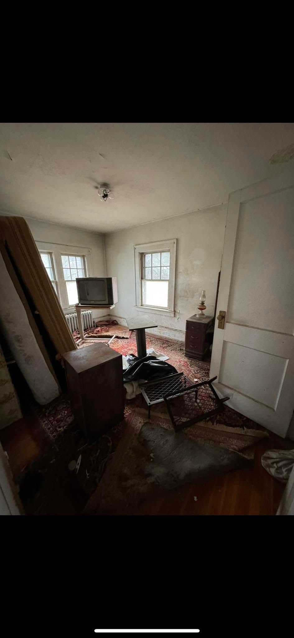A cluttered, sparsely furnished room with hardwood floors, a television on a stand, and debris scattered across the floor.