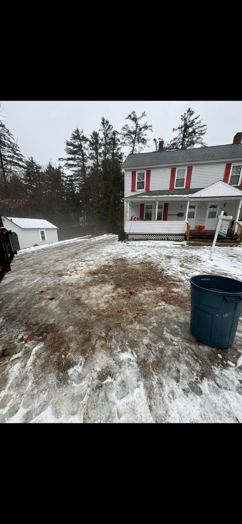 A snow-covered yard with patches of exposed dirt, a blue trash bin, and a two-story white house with red shutters.