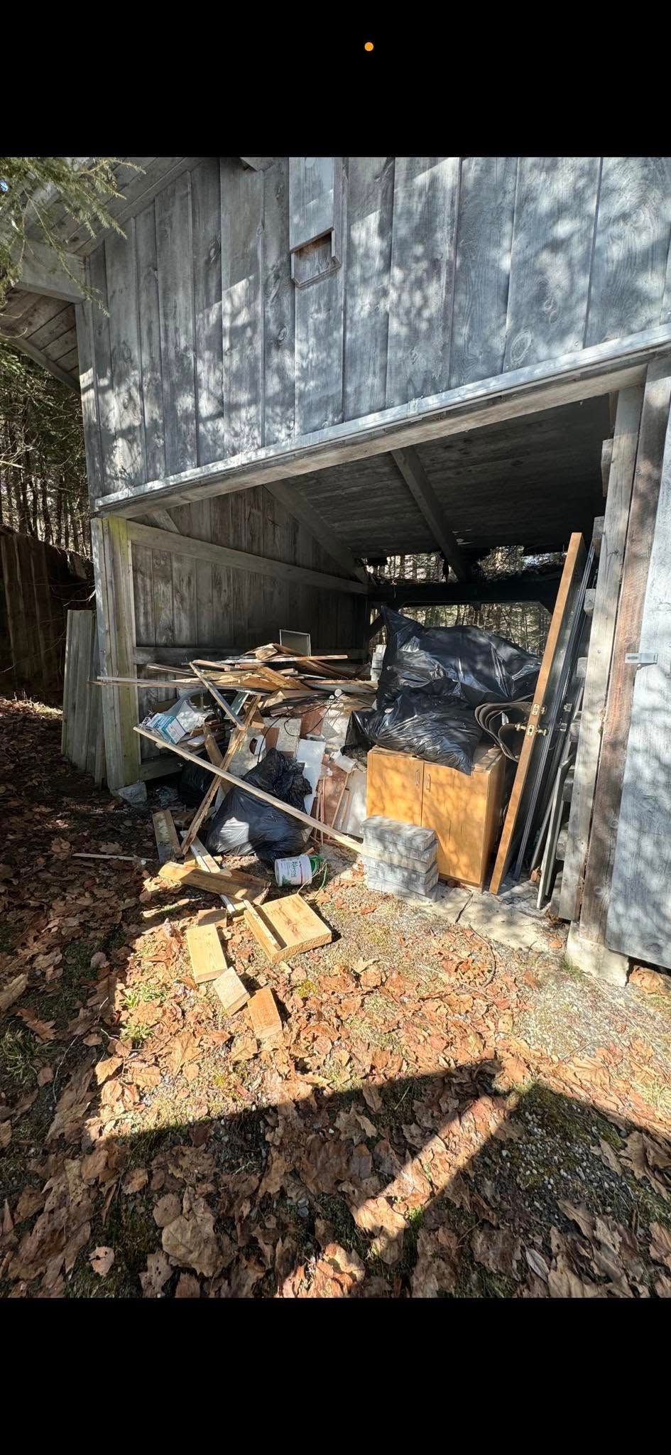 A cluttered, open-sided shed with debris, wood planks, and bags of trash piled underneath a corrugated metal roof.