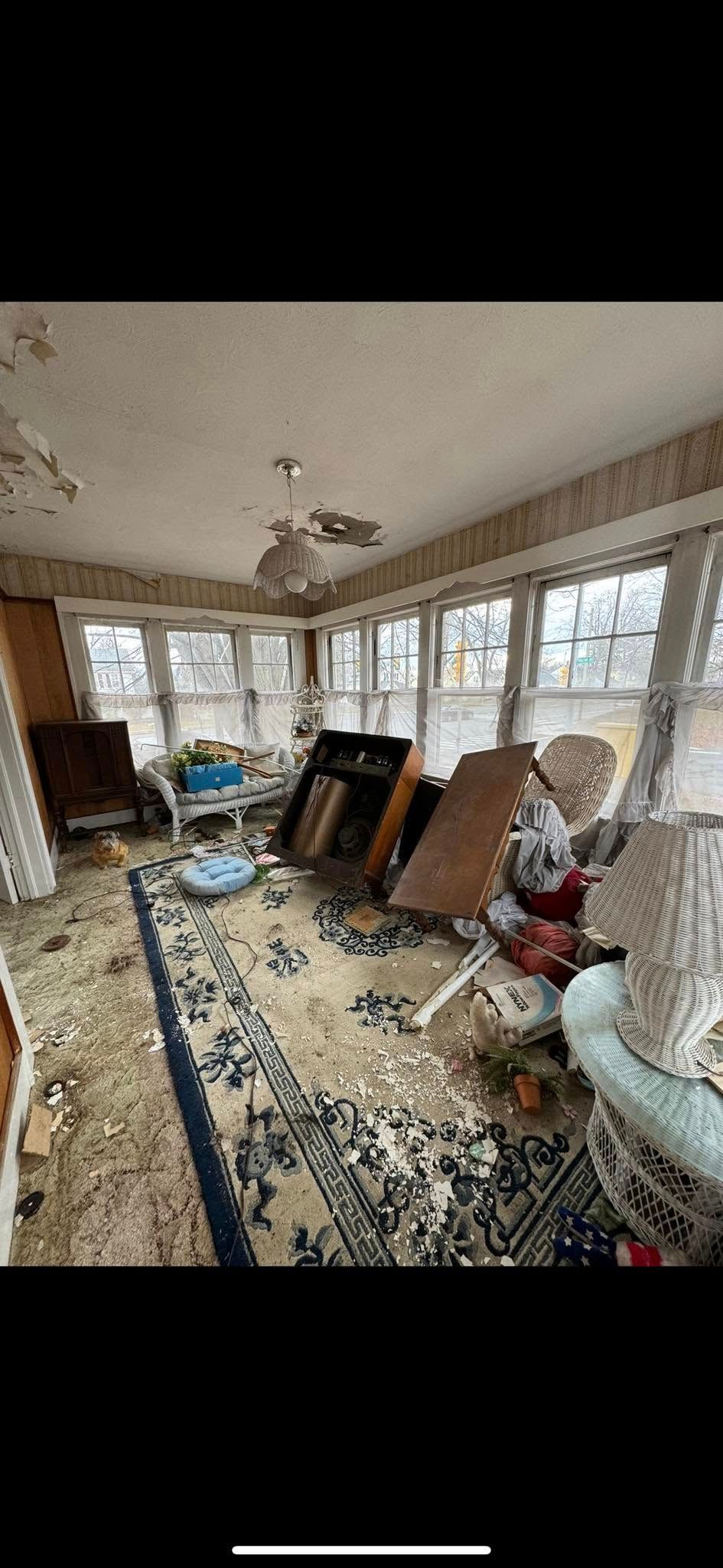 A sunroom filled with debris, scattered furniture, and a rug, showing signs of significant disrepair.