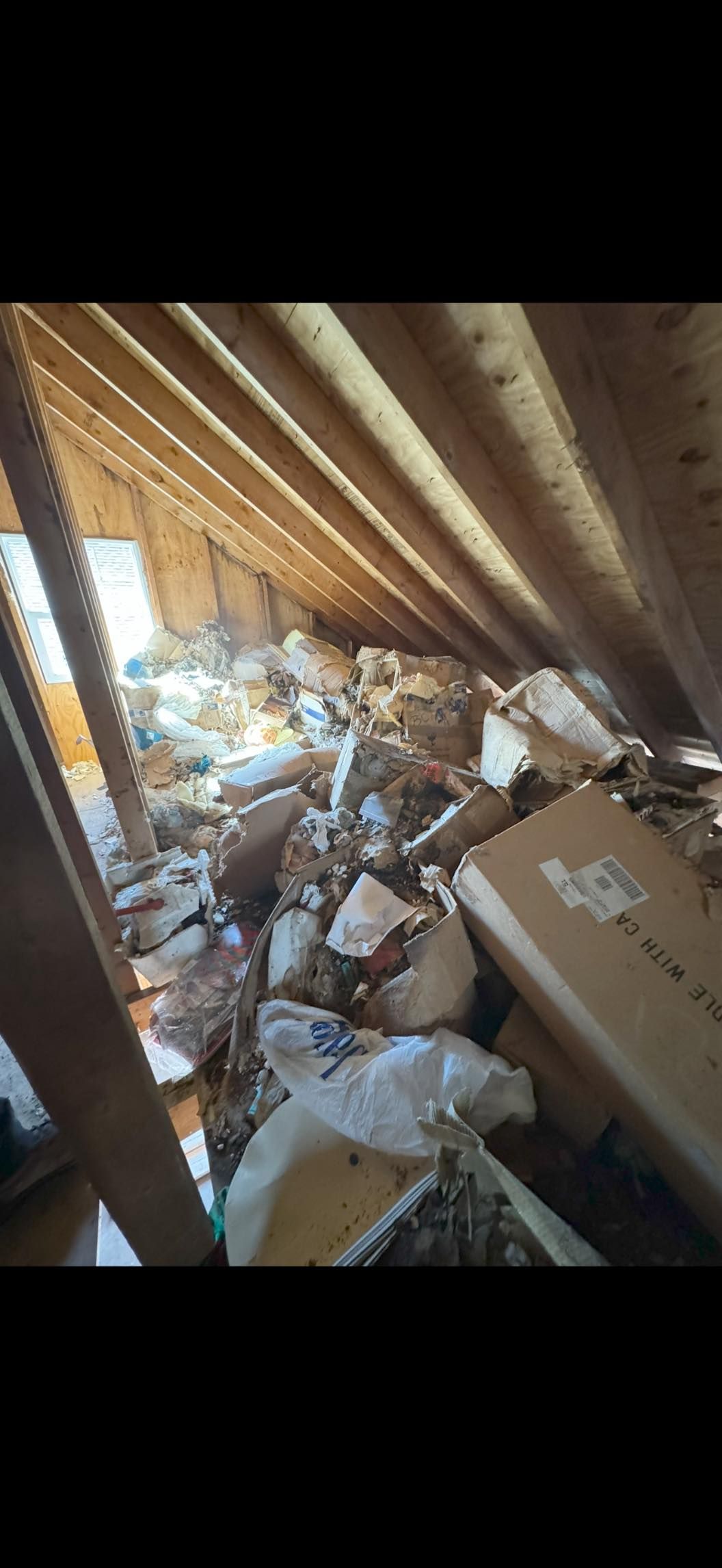 A cluttered attic space with exposed wooden roof rafters, filled with scattered cardboard boxes and debris.