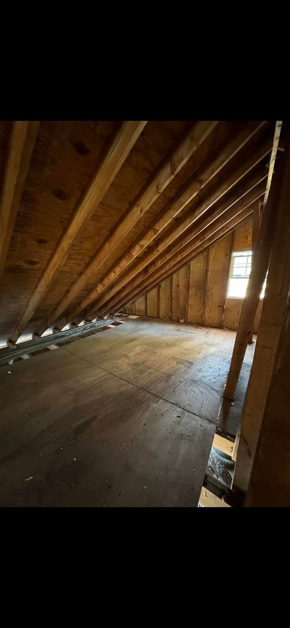 An unfinished attic with wooden rafters, subfloor, and a small window at the end, illuminated by natural light.