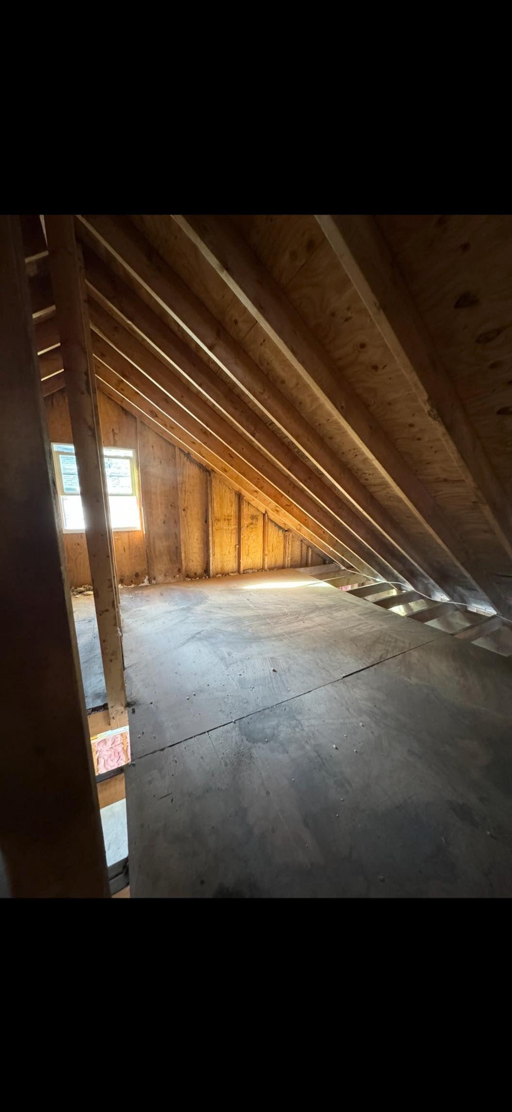 An attic interior with exposed wooden rafters, a plywood floor, and a window allowing light into the unfinished space.