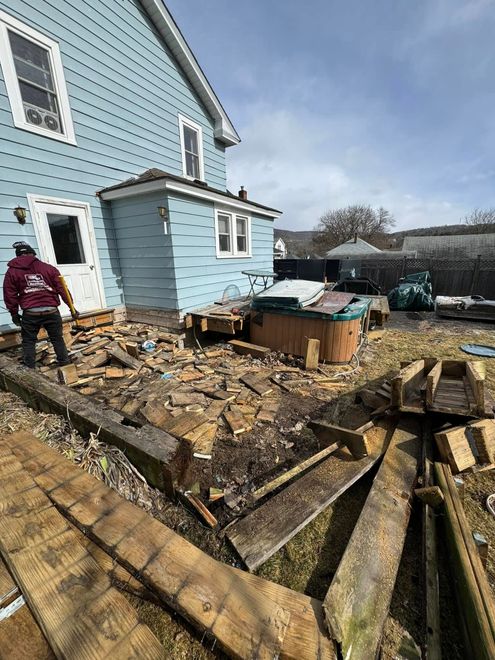 A person in a red jacket stands on a construction site outside a light blue house where a deck is being demolished.