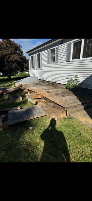 A person's shadow falls on a grassy yard next to a white house with a partially dismantled wooden deck.
