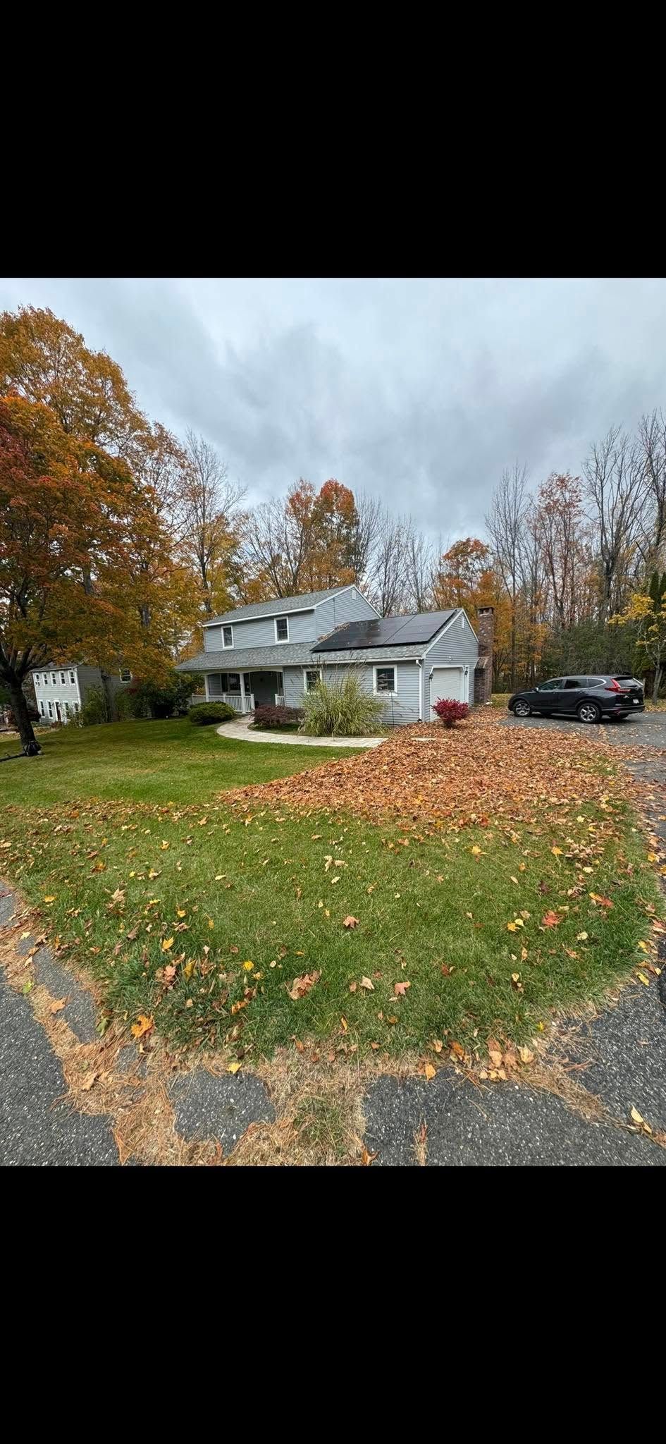 A two-story grey house surrounded by trees with autumn foliage and a lawn covered in fallen leaves under a cloudy sky.