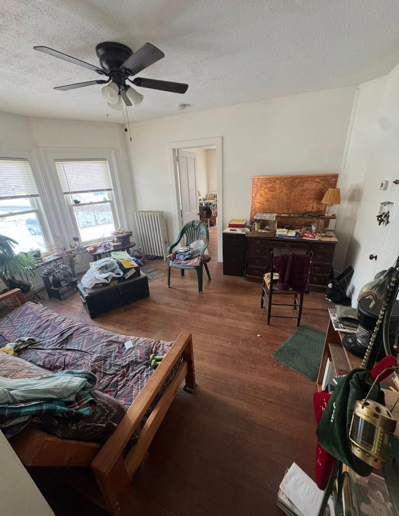 A sunlit room with a wooden bed frame, a desk, a chair, and a ceiling fan over hardwood floors.