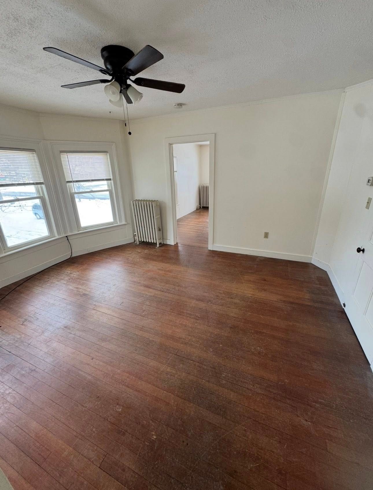 An empty room with dark wood flooring, beige walls, two windows with blinds, a ceiling fan, and a radiator.