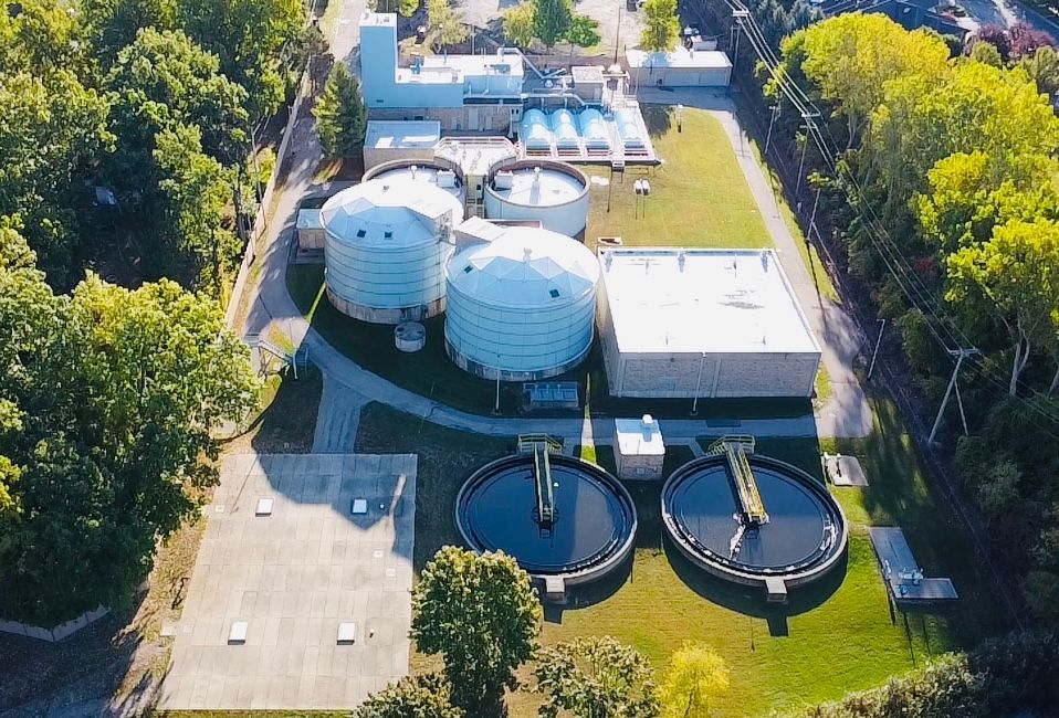 Aerial view of SWMUA wastewater treatment plant with large round tanks, buildings, and surrounding trees.